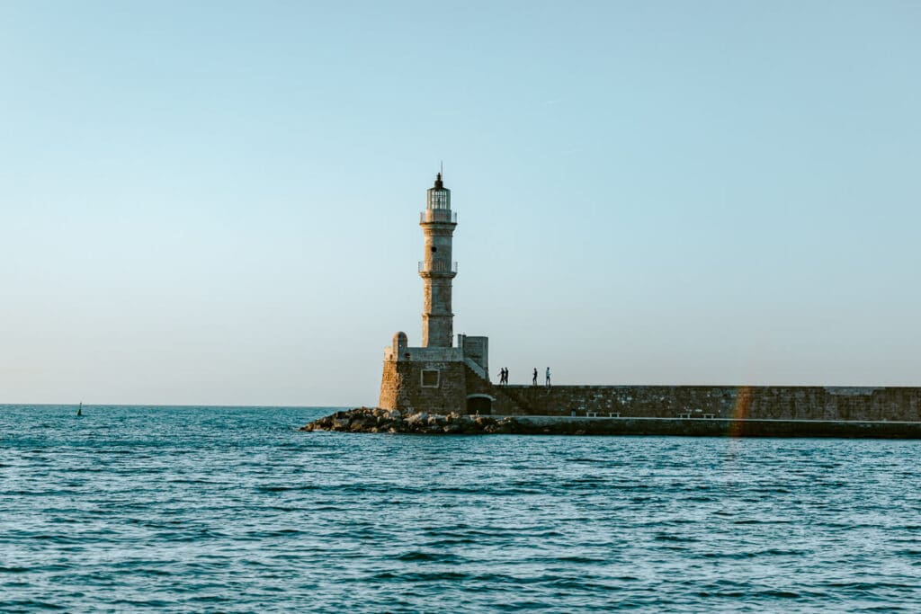 Stone lighthouse at the end of a harbor wall in Chania, Crete, with calm blue water in the foreground and a clear sky above.