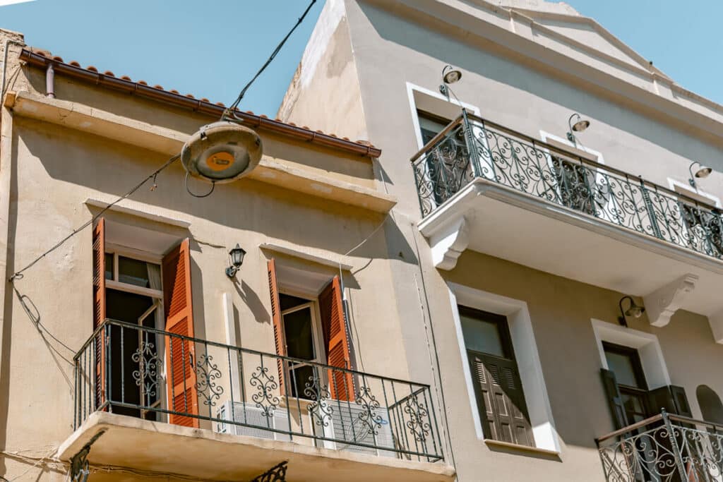 Greek island building with orange shutters, iron balconies, and pale walls under a clear sky, showing everyday architecture travelers may see while exploring local neighborhoods.