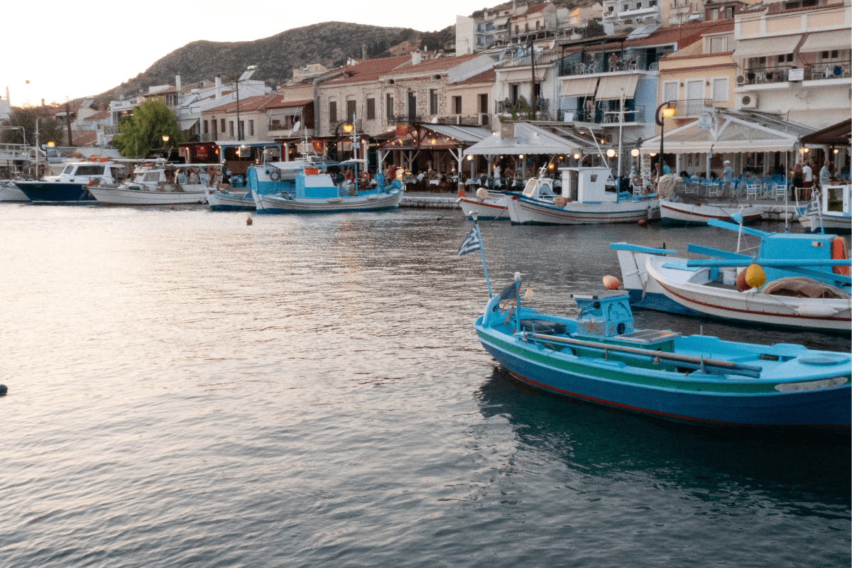 Blue fishing boats floating in a Greek harbor at sunset, with waterfront tavernas and hillside buildings in the background for a post answering is Greece expensive?