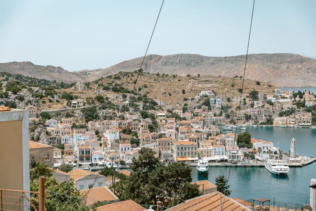 View over Symi Harbor from above, with pastel houses, boats, calm blue water, rocky hills, and the clock tower along the waterfront.