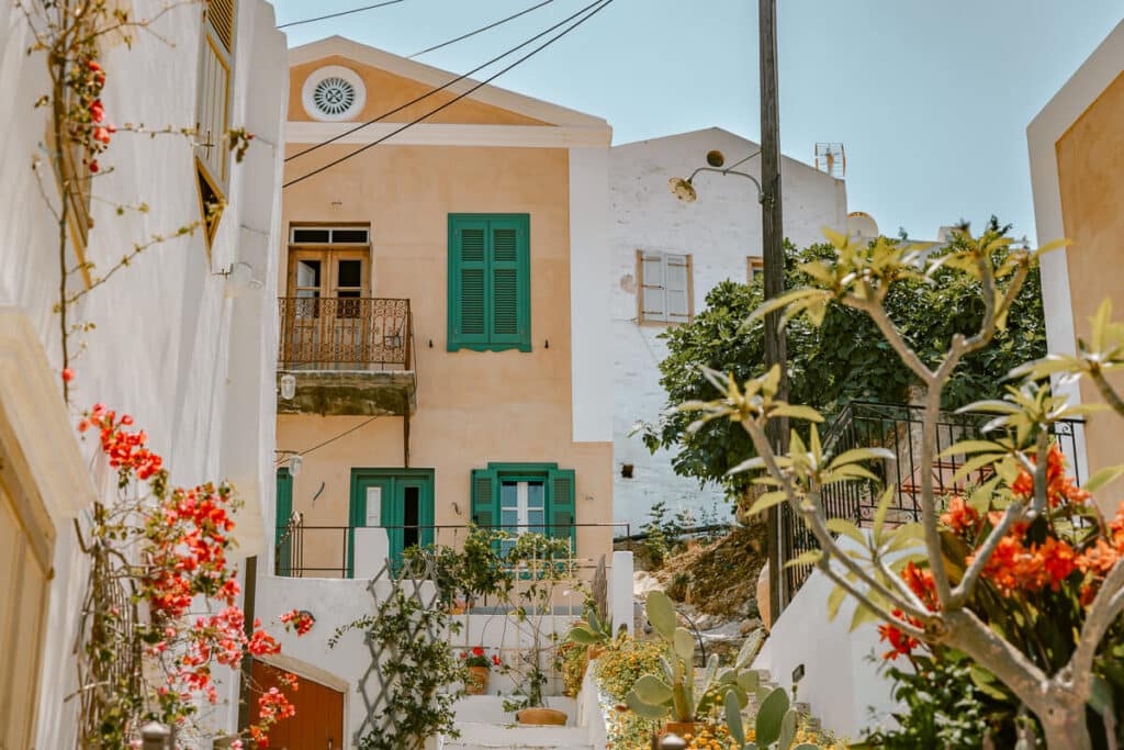Quiet hillside street in Symi with pastel homes, green shutters, white walls, flowering plants, and steps leading through Chorio.