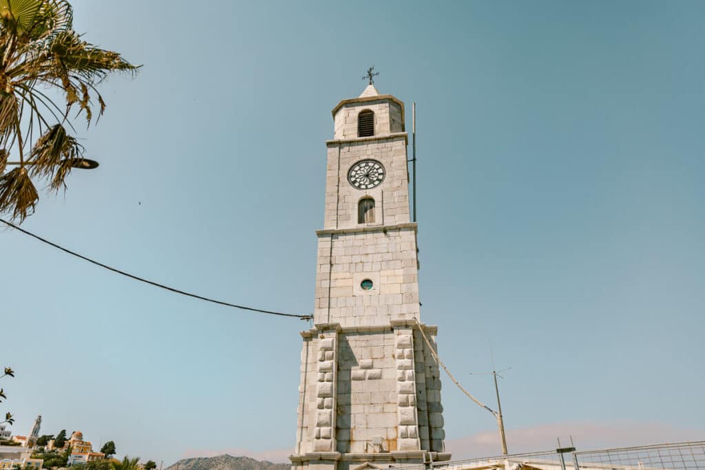 Stone clock tower near Symi Harbor with a blue sky, palm leaves, and colorful hillside buildings visible in the background.