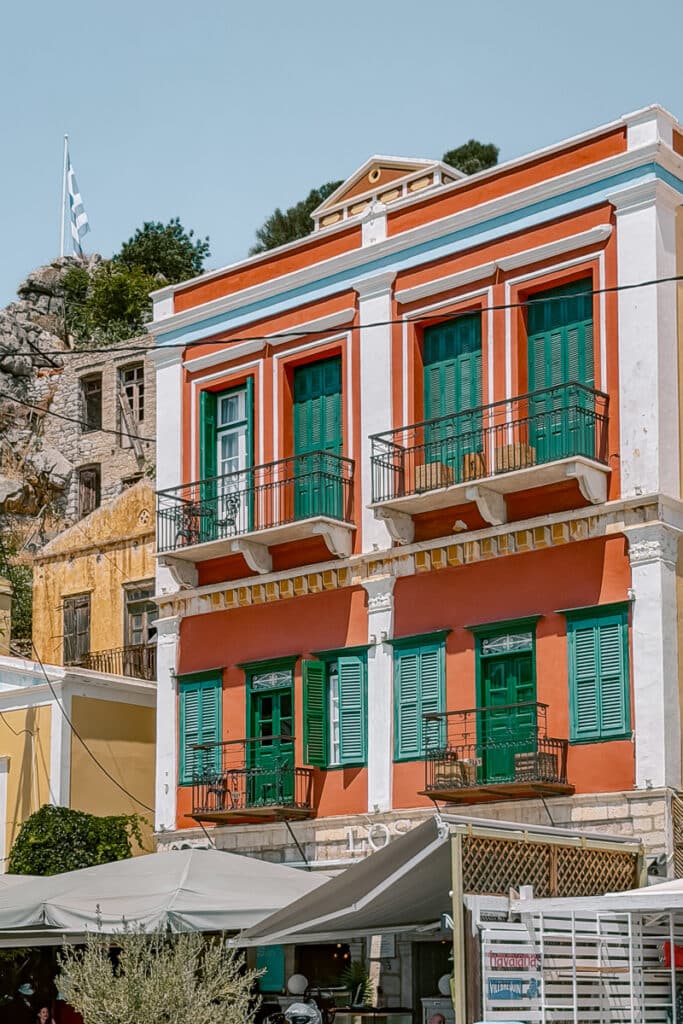 Close up of a bright red neoclassical building in Symi with green shutters, balconies, and cafe umbrellas below the colorful facade.