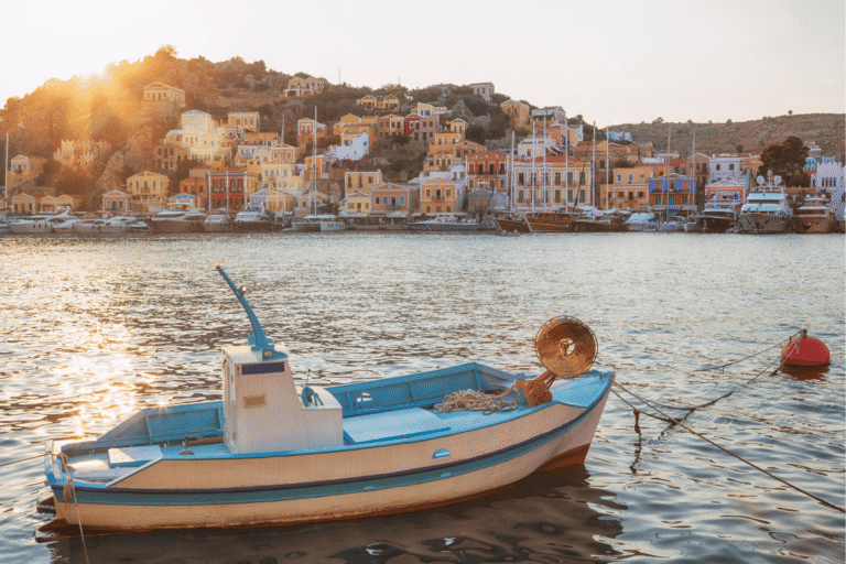Day trip to Symi from Rhodes with a small blue and white boat floating in Symi Harbor at sunset, colorful hillside houses, yachts, and warm golden light over the water.