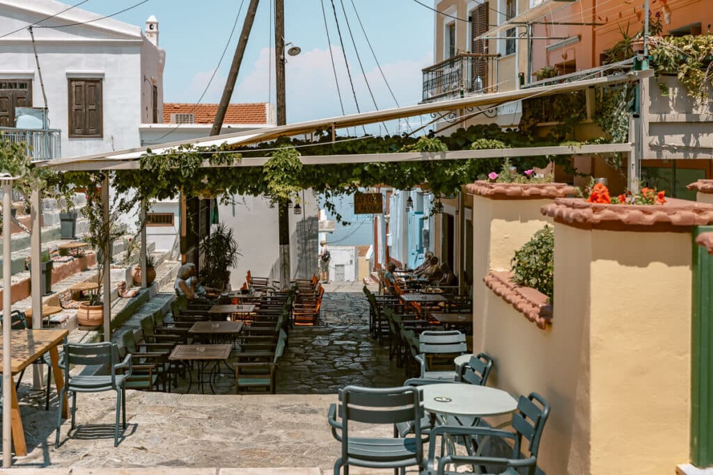 Outdoor restaurant seating on the stone steps of Kali Strata in Symi, with colorful buildings, shaded tables, and a view down toward the harbor.
