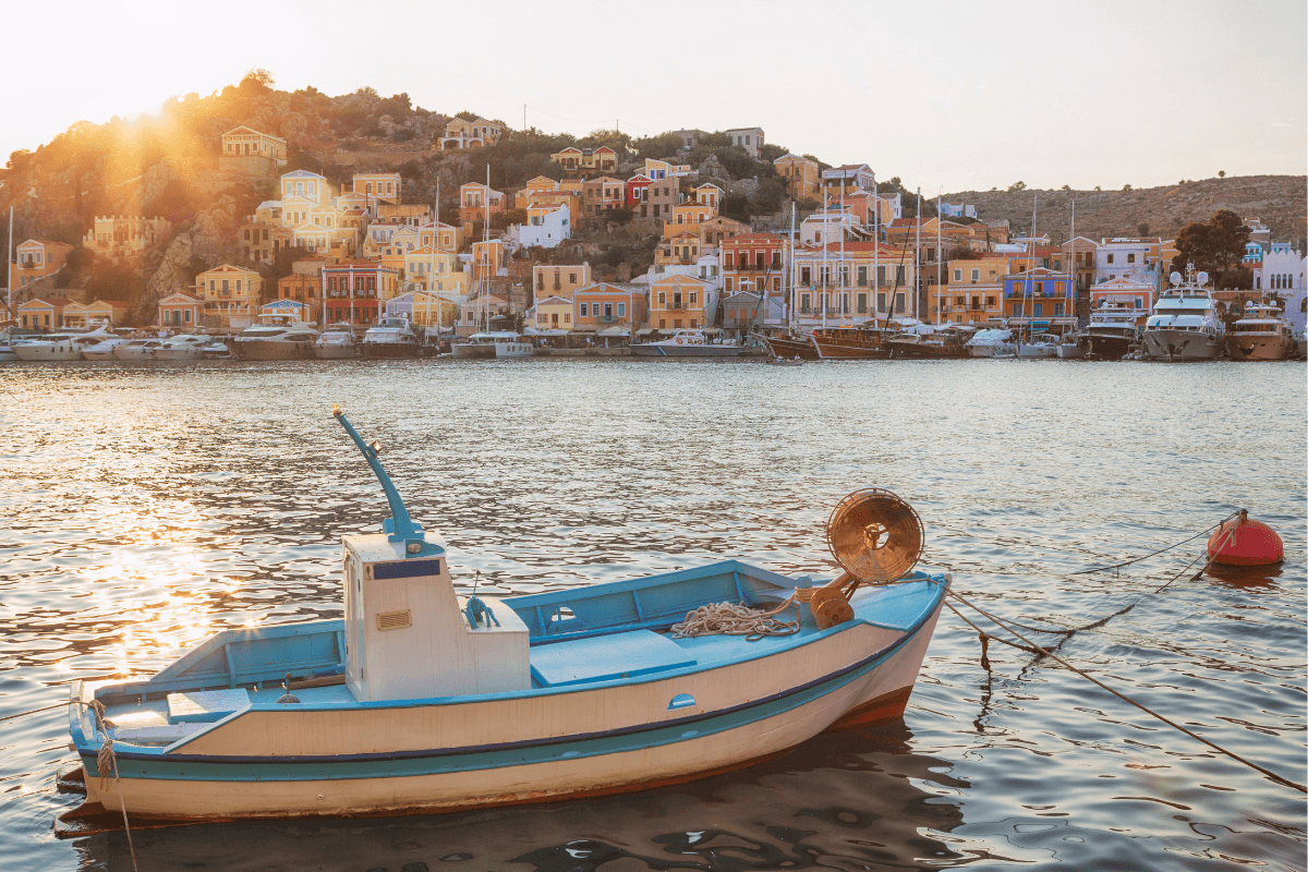 Day trip to Symi from Rhodes with a small blue and white boat floating in Symi Harbor at sunset, colorful hillside houses, yachts, and warm golden light over the water.