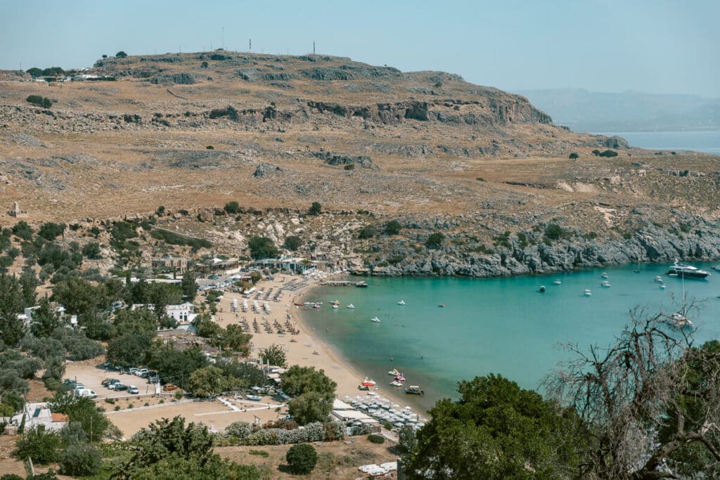 View over Lindos Beach in Rhodes, with turquoise water, boats, sandy shoreline, and dry rocky hills above the bay.