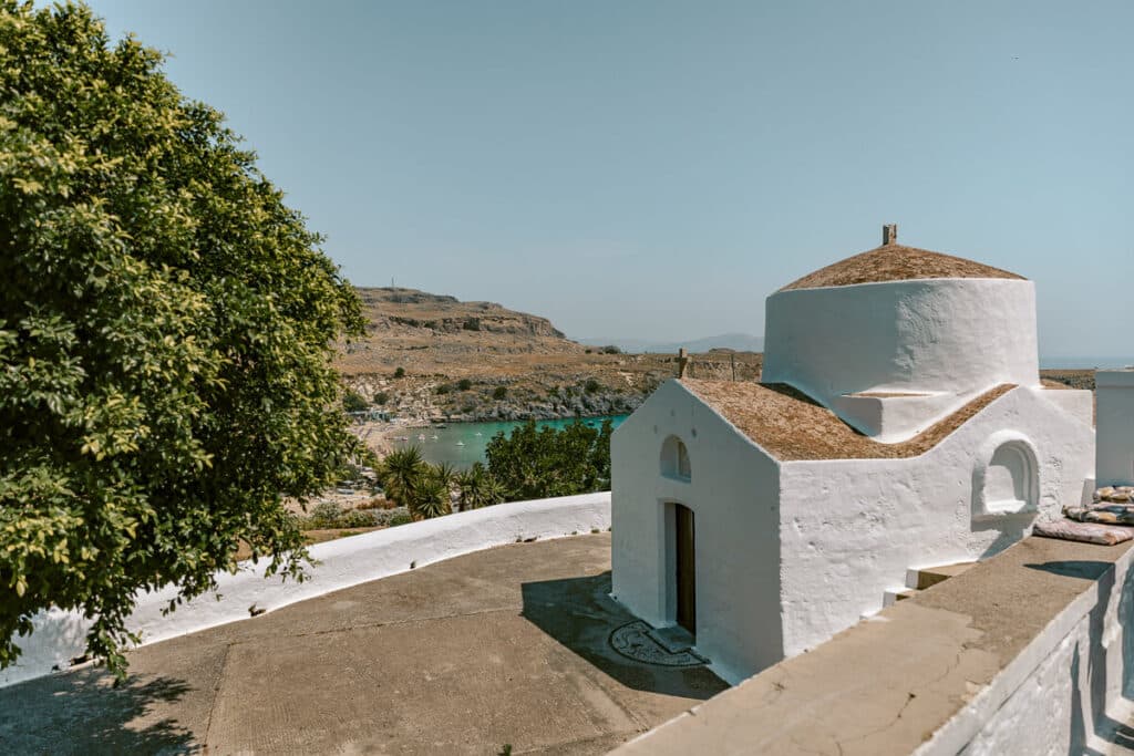 Small white chapel overlooking Lindos Beach in Rhodes, with turquoise water, rocky hills, and trees in the background.