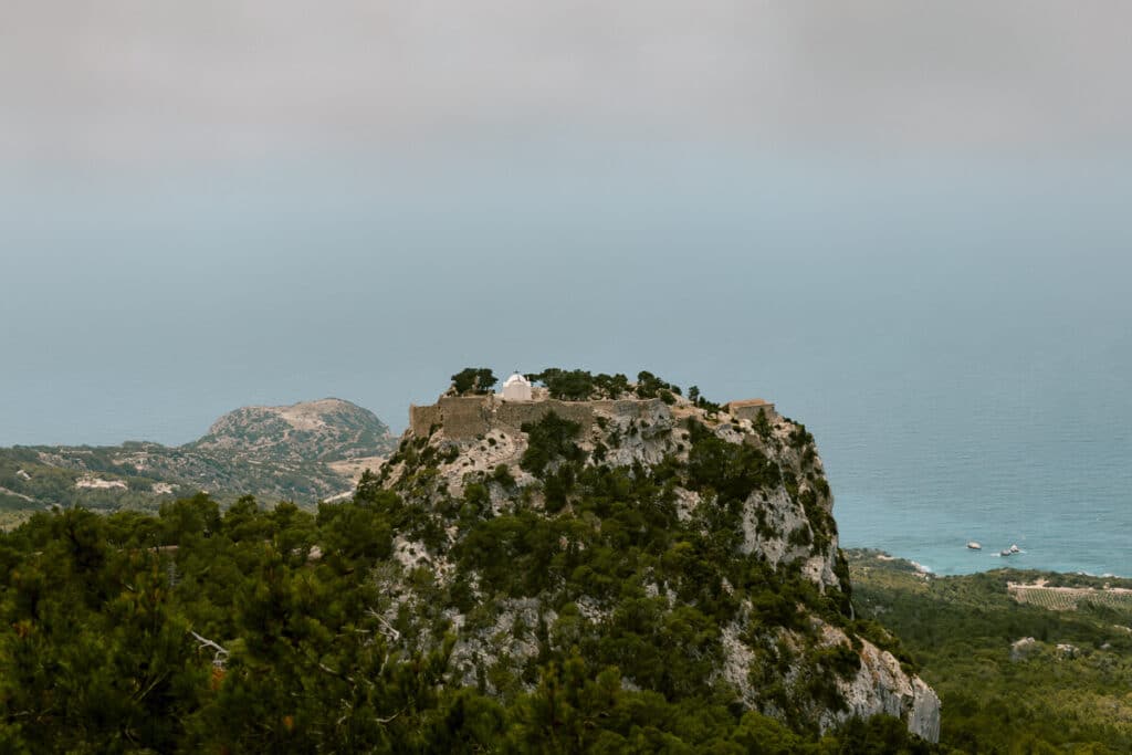 Castle of Monolithos on a rocky hilltop in Rhodes, with pine trees, sea views, and cliffs in the distance.