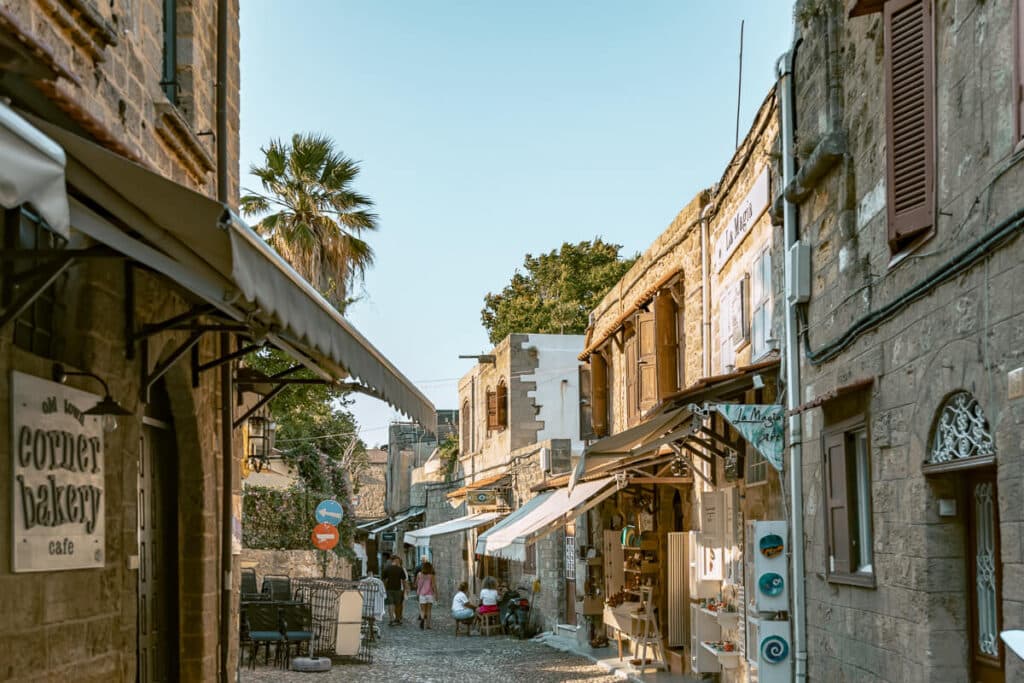 Cobblestone shopping street in Rhodes Old Town with stone buildings, awnings, cafe signs, and people walking in the distance.