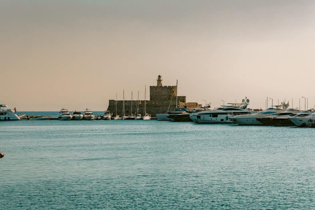 Mandraki Harbor in Rhodes with turquoise water, yachts, and the stone fortress of Saint Nicholas in the background.