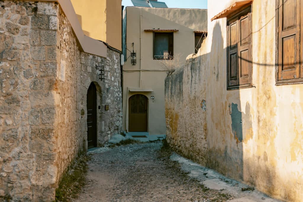 Narrow cobblestone street in Rhodes Old Town lined with old stone buildings, wooden shutters, and sunlit weathered walls.