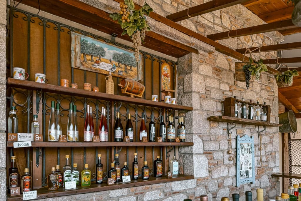 Wine, souma, and local bottles displayed on wooden shelves inside a stone winery or shop in Rhodes, with hanging grapes and rustic decor.