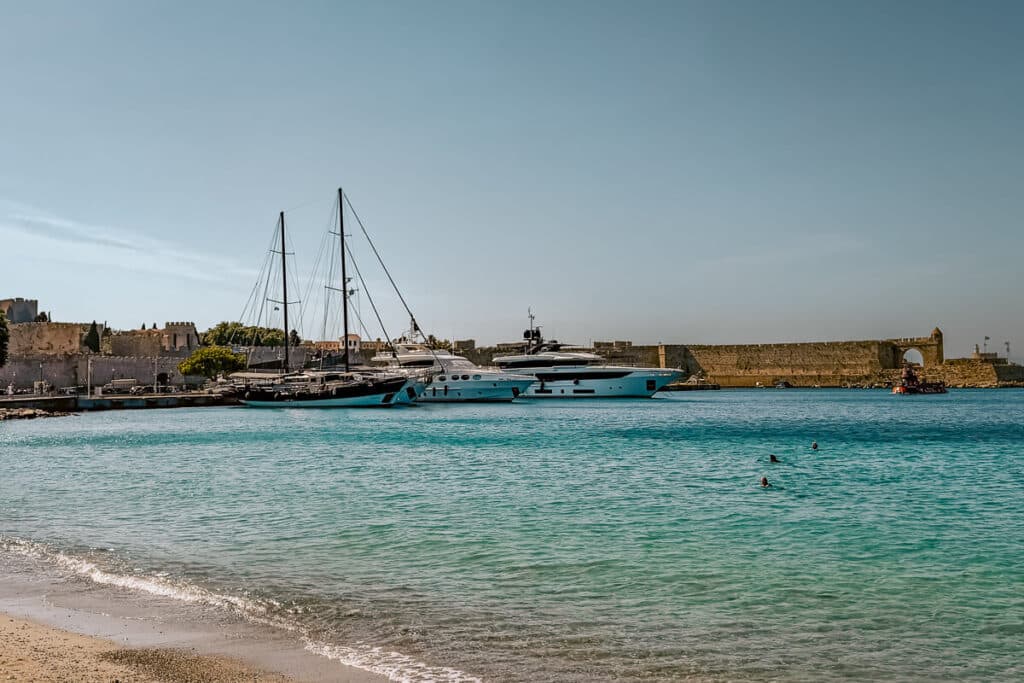 Clear turquoise water at a beach in Rhodes Town with swimmers, yachts, and medieval harbor walls in the background.