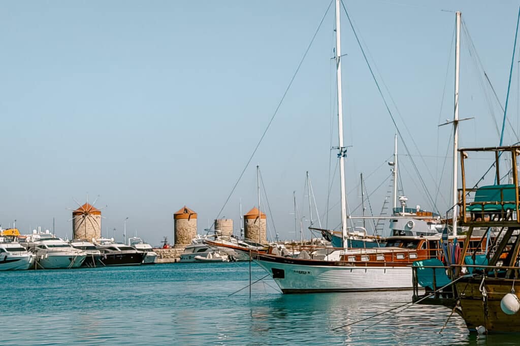 Traditional windmills at Mandraki Harbor in Rhodes, with sailboats, yachts, and calm turquoise water in the foreground.