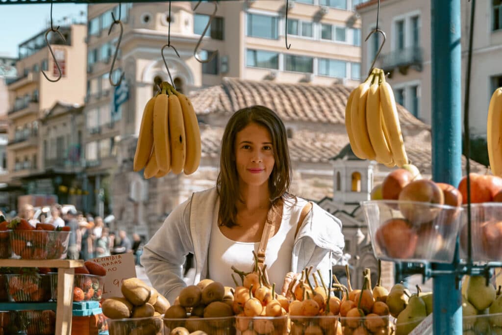 Me standing at a fruit stand in Monastiraki, Athens, surrounded by bananas, apples, and pears, with city buildings and a small church behind me.