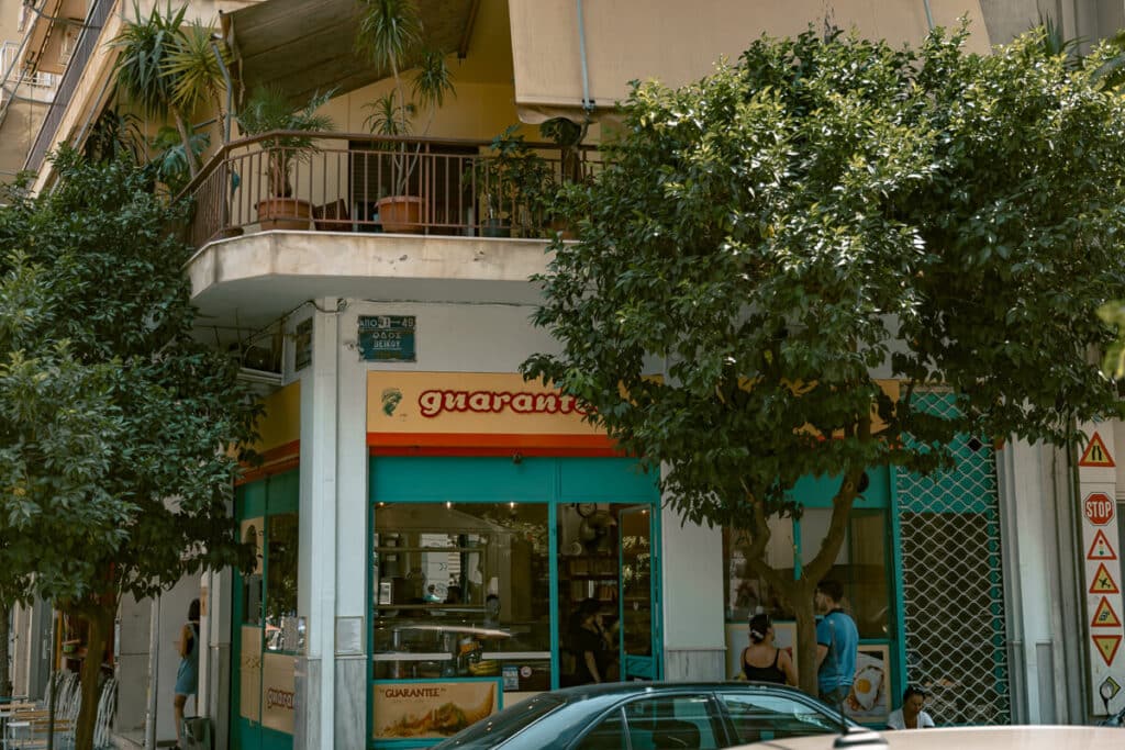 Street corner in Koukaki, Athens, with leafy trees, apartment balconies, and a colorful cafe storefront, showing the neighborhood’s local feel near the city center.