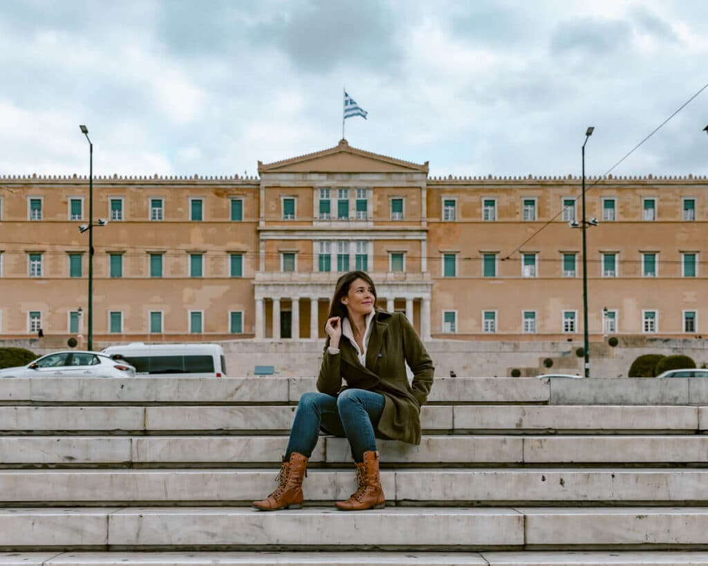 Me sitting on marble steps in Syntagma Square, with the Hellenic Parliament building and Greek flag behind me in central Athens.