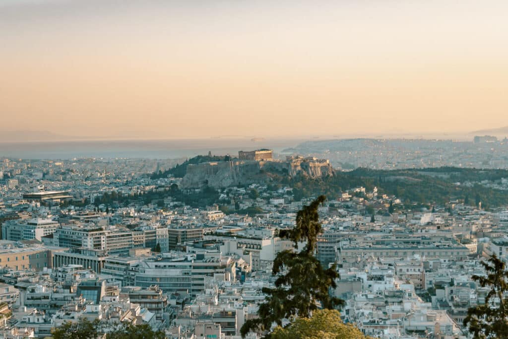 View from Prasini Tenta in Kolonaki, Athens, overlooking the city rooftops, the Acropolis, and the sea in the distance at sunset.