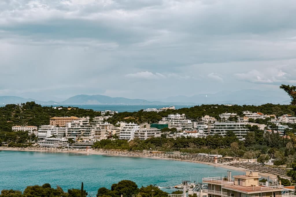 View over Vouliagmeni on the Athens Riviera, with turquoise water, beachfront hotels, tree-covered coastline, and mountains in the distance under a cloudy sky.