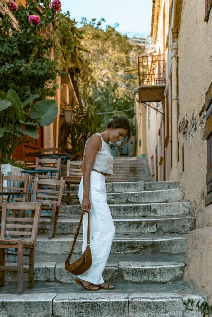 Me standing on stone steps in Plaka, Athens, surrounded by old buildings, greenery, cafe chairs, and narrow historic streets near the Acropolis.