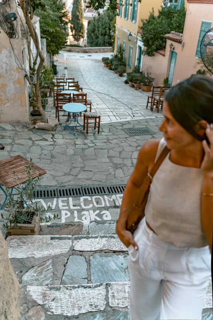 Me standing on stone steps with “Welcome Plaka” painted below, overlooking cafe tables, colorful buildings, and a quiet street in Plaka, Athens.