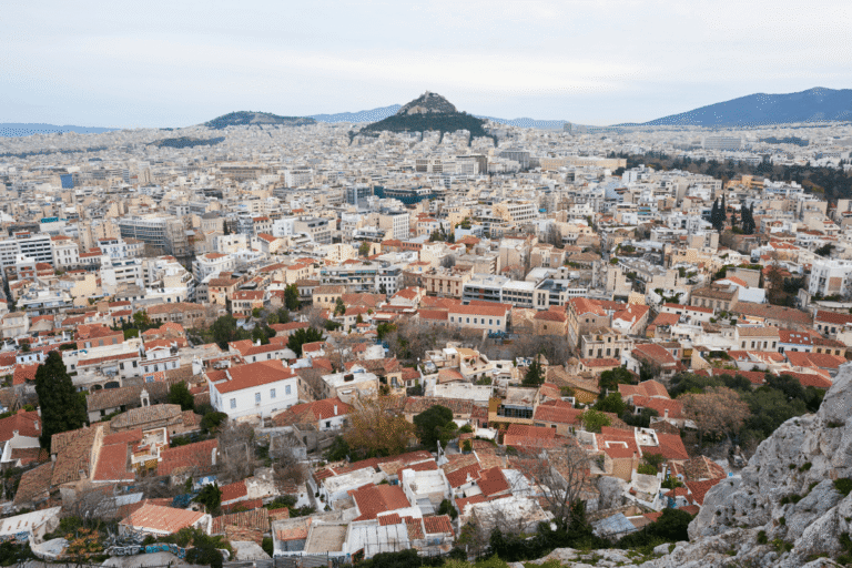 Panoramic view over Athens from above, with red rooftops in the foreground, dense city buildings stretching toward Lycabettus Hill, and mountains in the distance, for a where to stay in Athens guide.