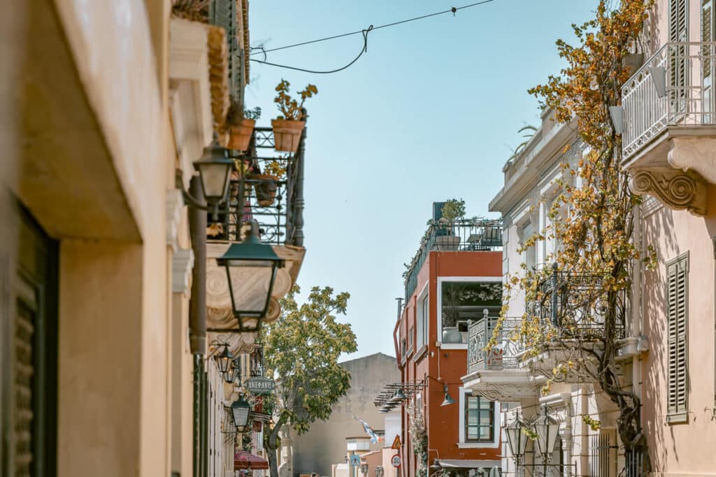 Narrow street in Plaka, Athens, lined with neoclassical buildings, balconies, shutters, street lamps, and climbing greenery on a sunny day.