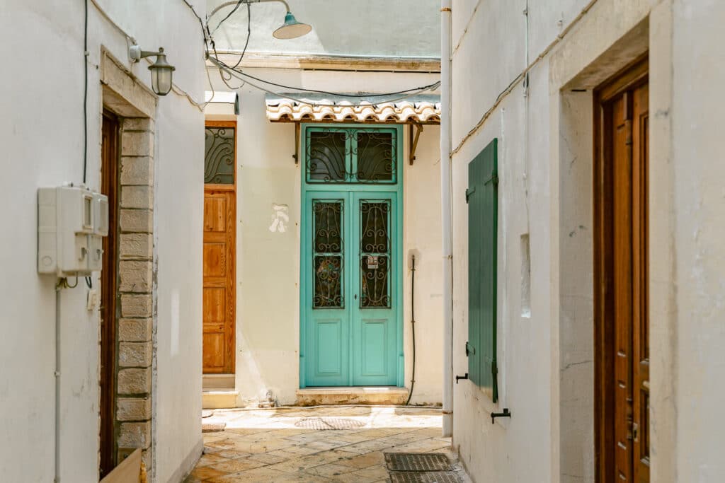Narrow street in Paxos with whitewashed walls, wooden doors, green shutters, and a turquoise door in a quiet village alley.