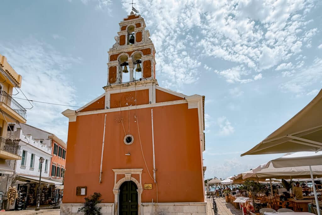 Orange church with a bell tower in Gaios, Paxos, surrounded by outdoor restaurant tables and village buildings under a cloudy blue sky.