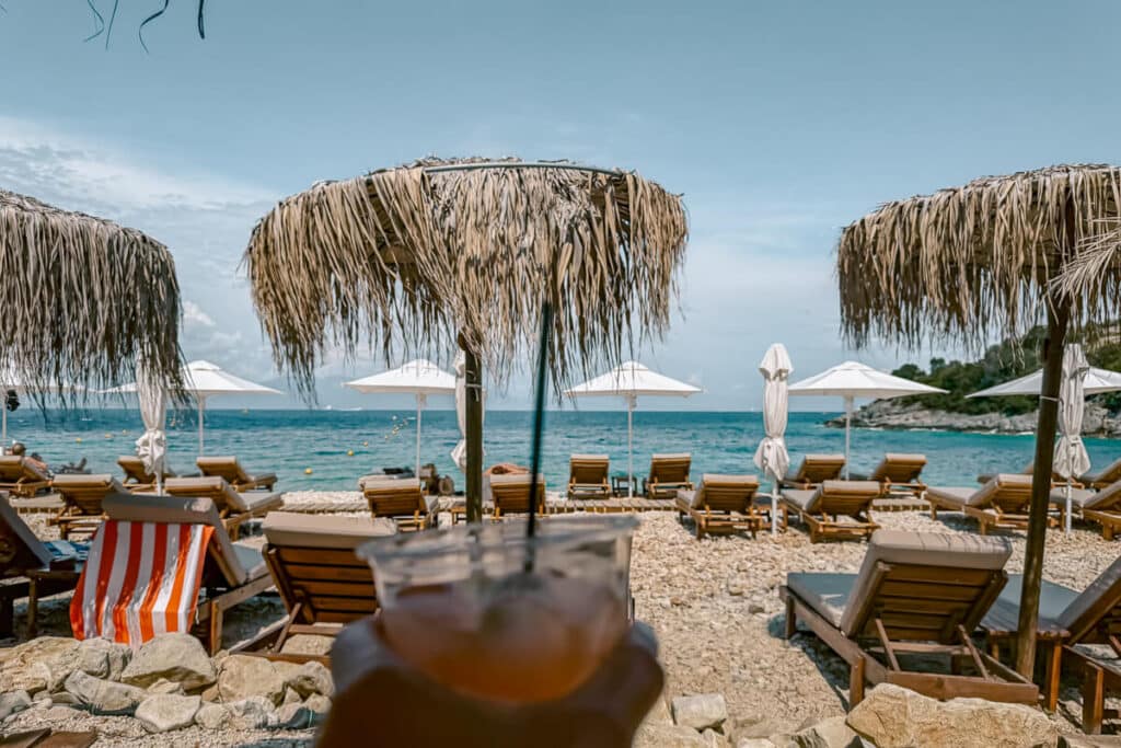Pebbly beach in Paxos with sunbeds, straw umbrellas, white umbrellas, turquoise water, and a drink held in the foreground.