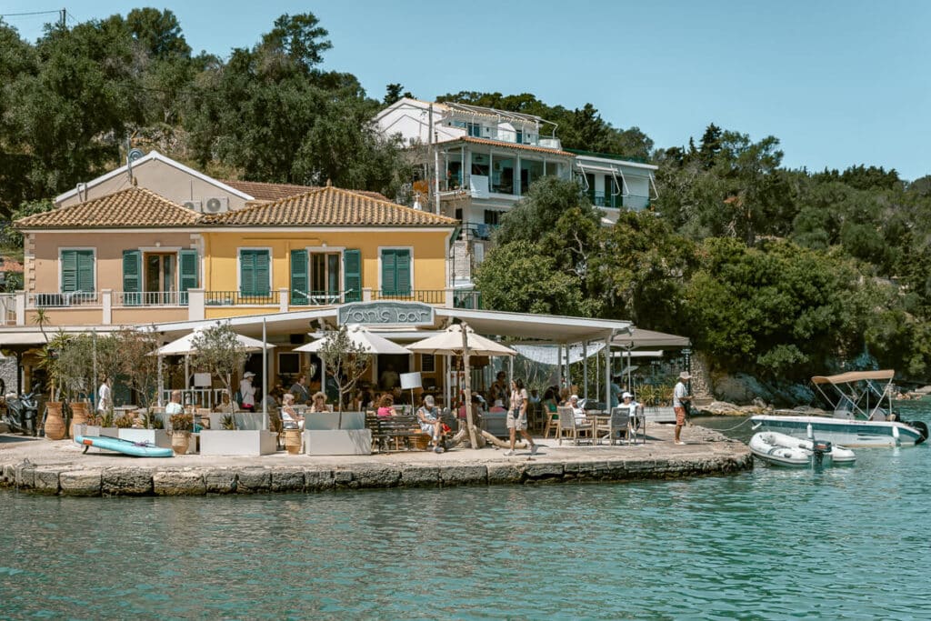 Waterfront restaurant in Paxos with outdoor tables, pastel buildings, small boats, and clear turquoise water along the harbor.