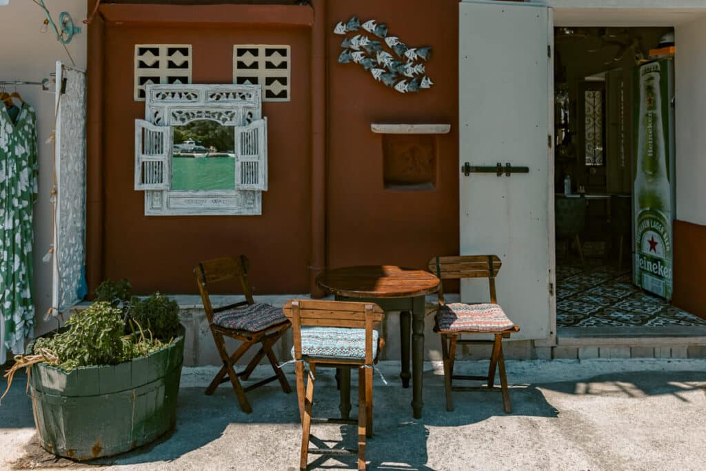 Small outdoor cafe table and chairs outside a shop in Paxos with a decorative mirror, plants, and an open doorway.