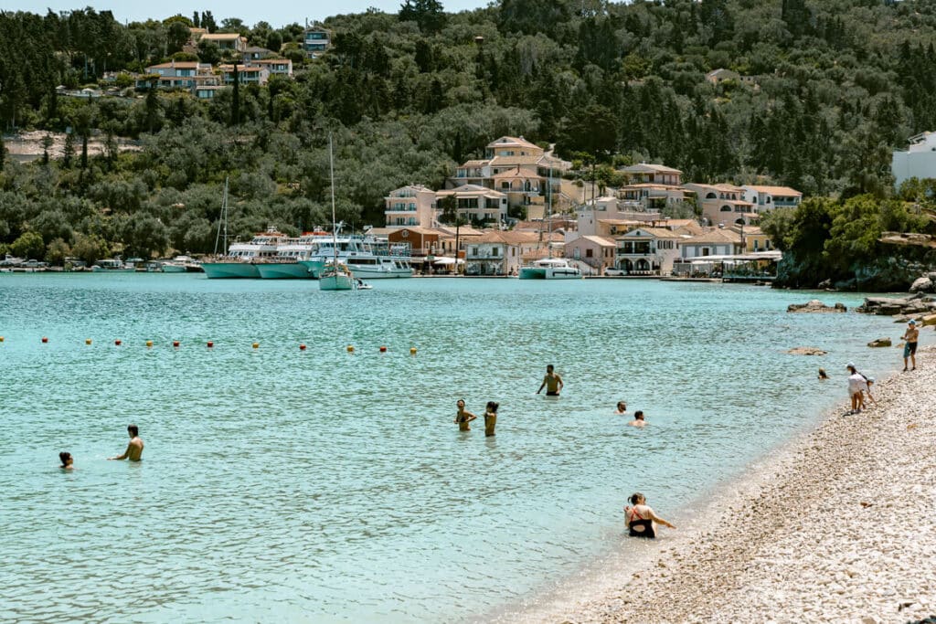 People swimming in pale turquoise water beside a pebbly beach in Paxos with village buildings and boats in the background.