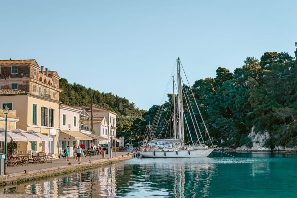 Waterfront village in Paxos with pastel buildings, outdoor cafe tables, a sailboat, and clear turquoise harbor water.