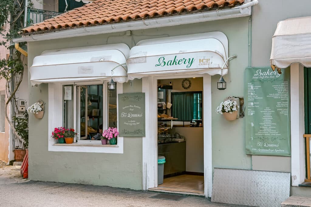 Exterior of Bakery To Spitiko in Paxos with a pale green storefront, white awnings, flower pots, and the bakery entrance open.