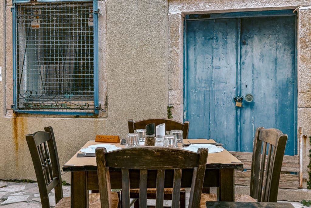 Outdoor restaurant table set for a meal in Paxos with wooden chairs, glassware, a blue door, and a stone wall in the background.