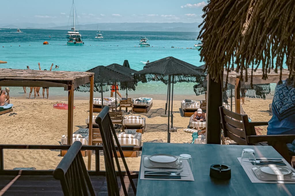 Beach restaurant table overlooking white sand, sunbeds, umbrellas, boats, and turquoise water at Paralia Vrika on Antipaxos Island.