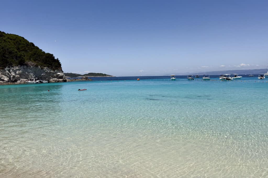 Clear shallow water and white sand at Paralia Vrika on Antipaxos Island with boats floating offshore.