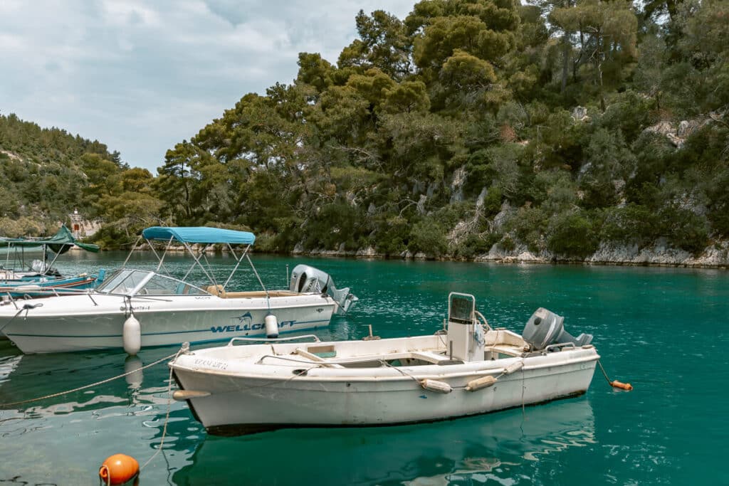 Small boats moored in clear turquoise water beside a rocky, tree-covered shoreline in Paxos.