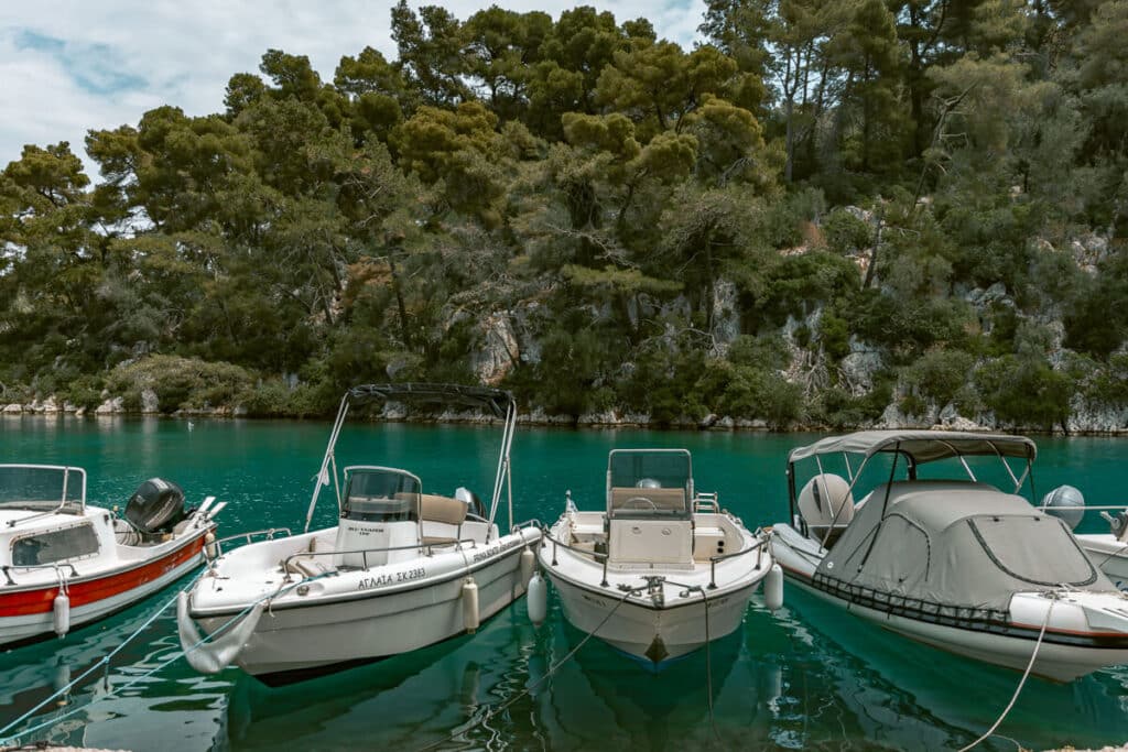 Small boats floating in clear turquoise water near a rocky, tree-covered shoreline in Paxos.