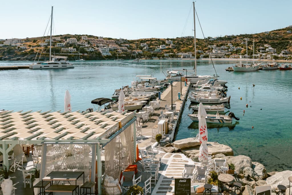 Batsi harbor in Andros with small boats, clear blue water, seaside cafes, and white houses on the hillside.