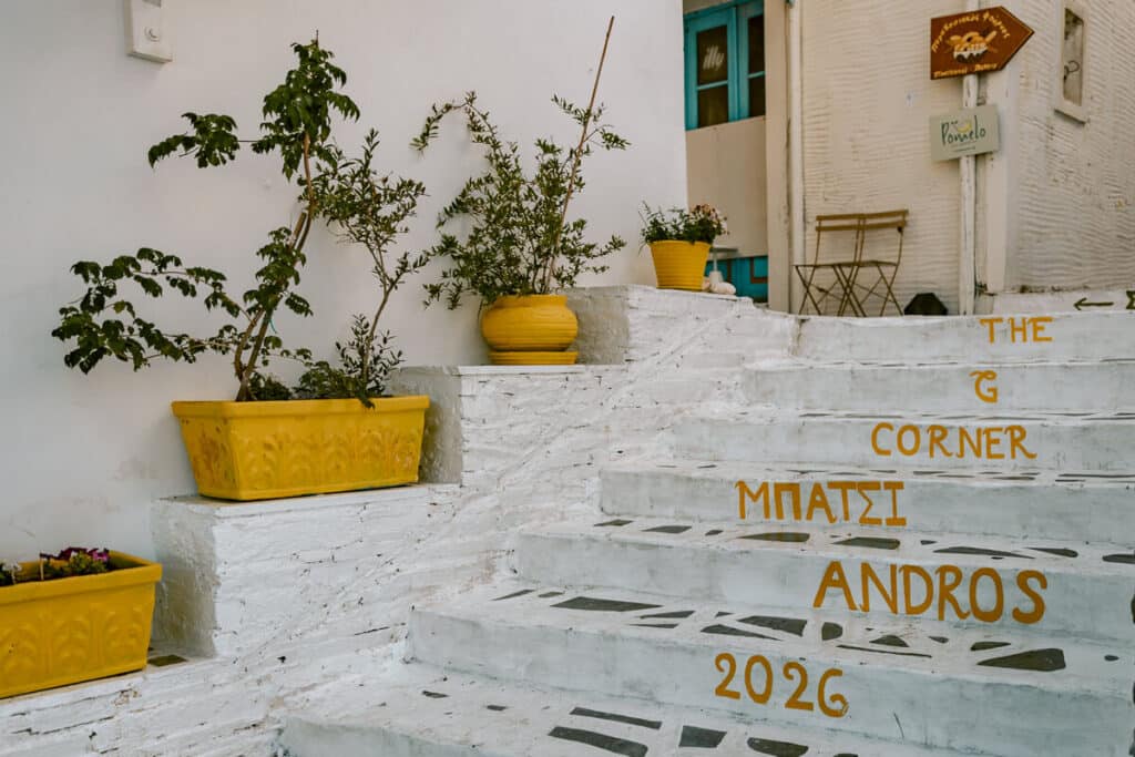 White steps in Batsi, Andros, with yellow planters, Greek writing, and signs for nearby cafes and shops.
