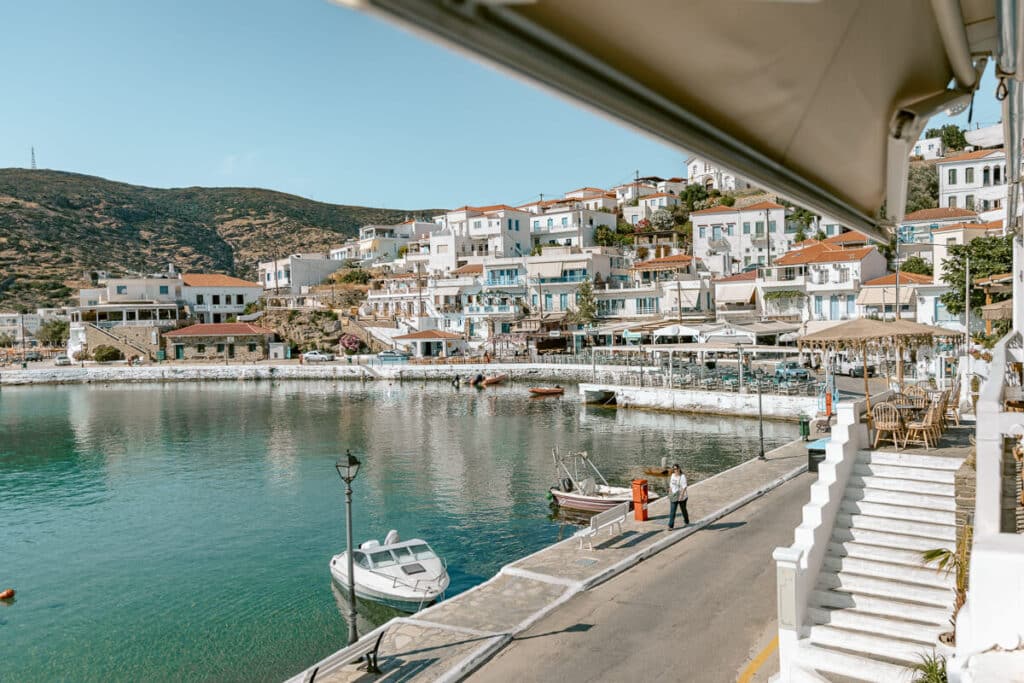 View of Batsi harbor in Andros, Greece, with calm turquoise water, small boats, white houses, and seaside cafes along the waterfront.