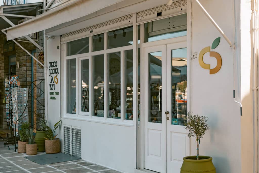 Storefront of παμπιλονι in Andros, Greece, with white doors, large windows, potted plants, and local gifts displayed inside.