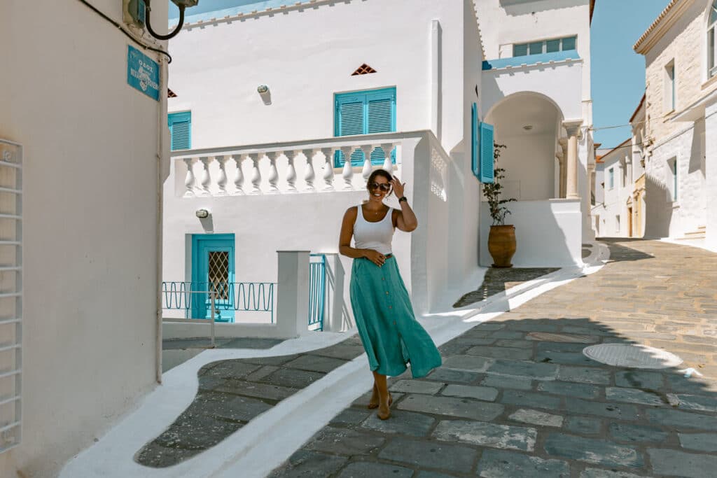 A woman walking through the whitewashed streets of Chora, Andros, surrounded by blue shutters, stone paths, and traditional Cycladic buildings.
