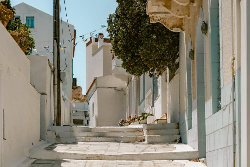 White stone steps and narrow streets in Chora, Andros, with whitewashed buildings, blue shutters, a leafy tree, and Greek flags overhead.