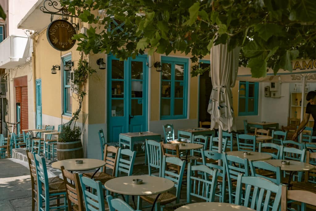 Outdoor seating at Νοσταλγία 1950 in Chora, Andros, with turquoise chairs, shaded tables, blue doors, and leafy trees overhead.