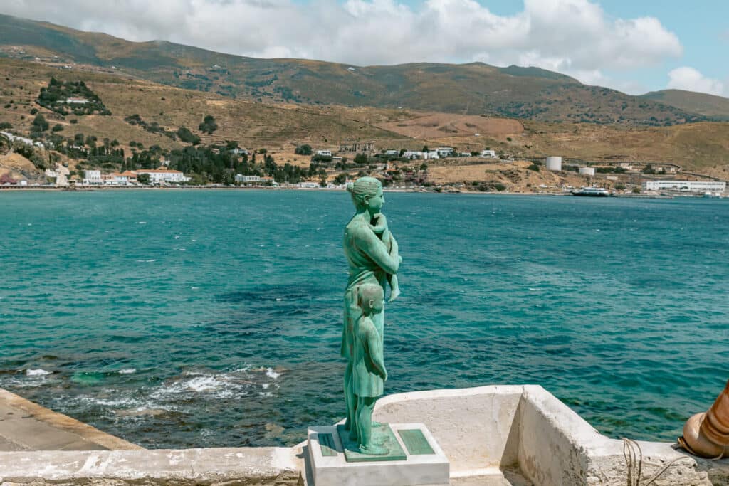 Bronze statue of a woman and child overlooking the turquoise sea and hills in Andros, Greece.