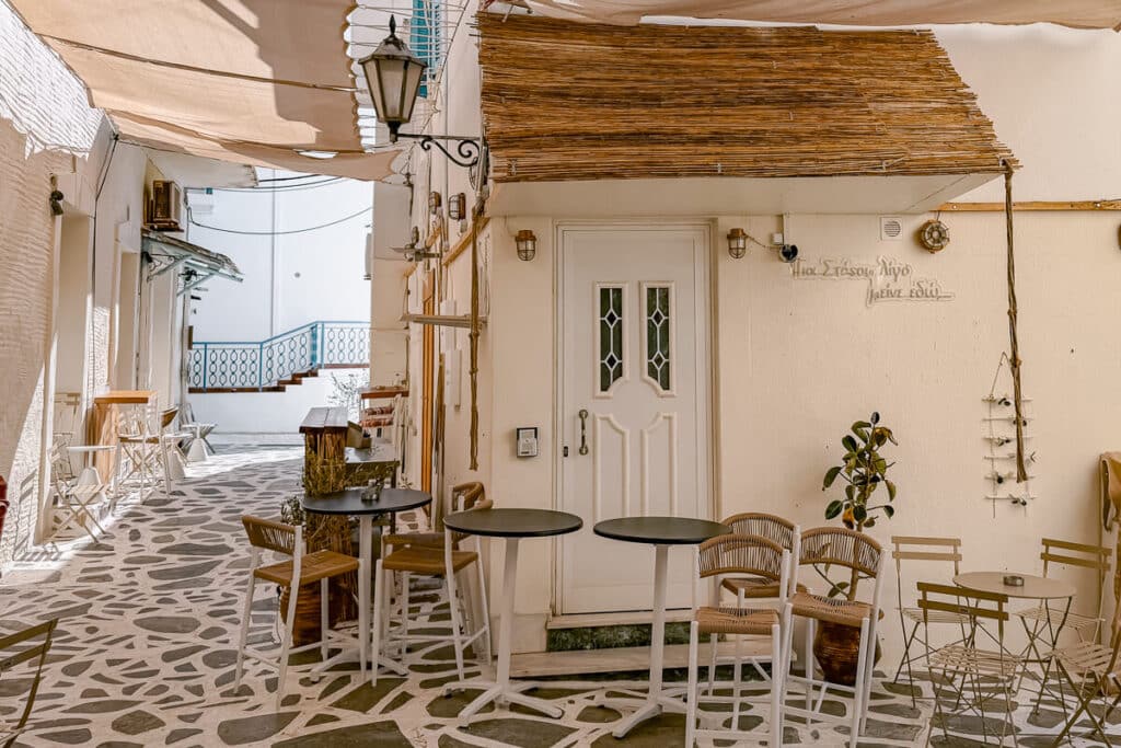 Outdoor cafe tables and chairs on a narrow stone street in Chora, Andros, with white buildings, a lantern, and shaded awnings.